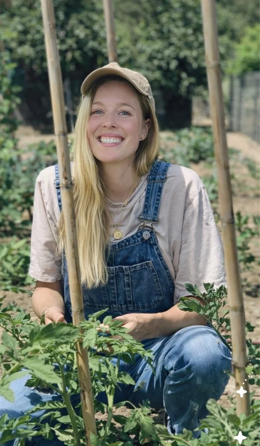 Benny the nutritionist in the garden, wearing a beige cap, beige t-shirt, and denim overalls. Benny is smiling while sitting in a garden among green plants and tomato plants supported by bamboo stakes.
