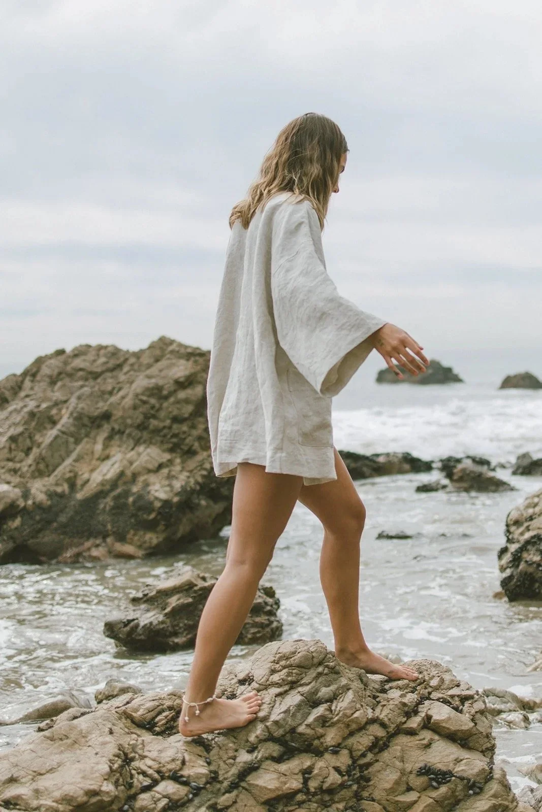 Woman in a white robe standing barefoot on rocks by the beach, looking down, with the ocean and cloudy sky in the background.