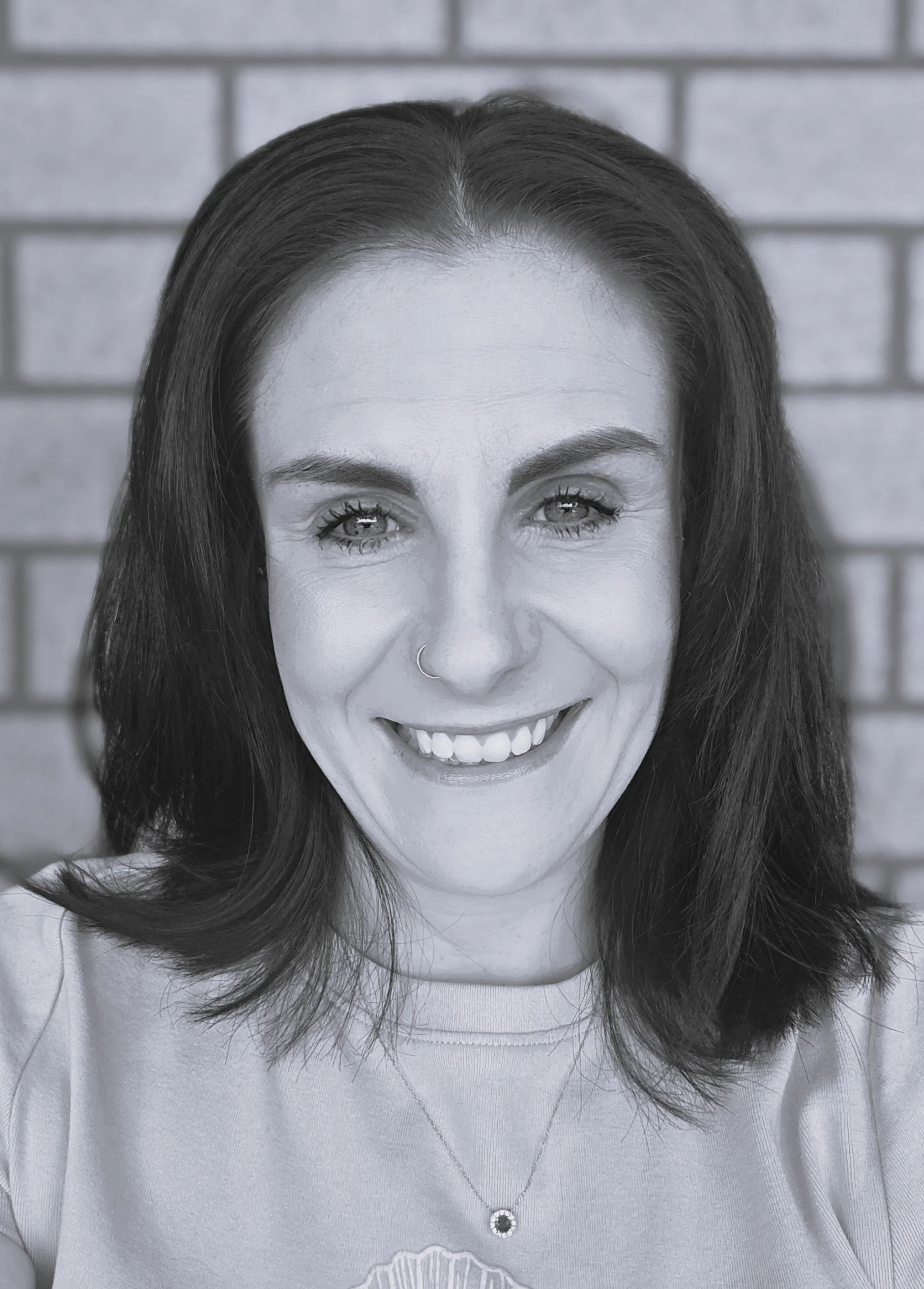 A smiling woman with dark hair, wearing a nose ring and a necklace, standing in front of a brick wall.