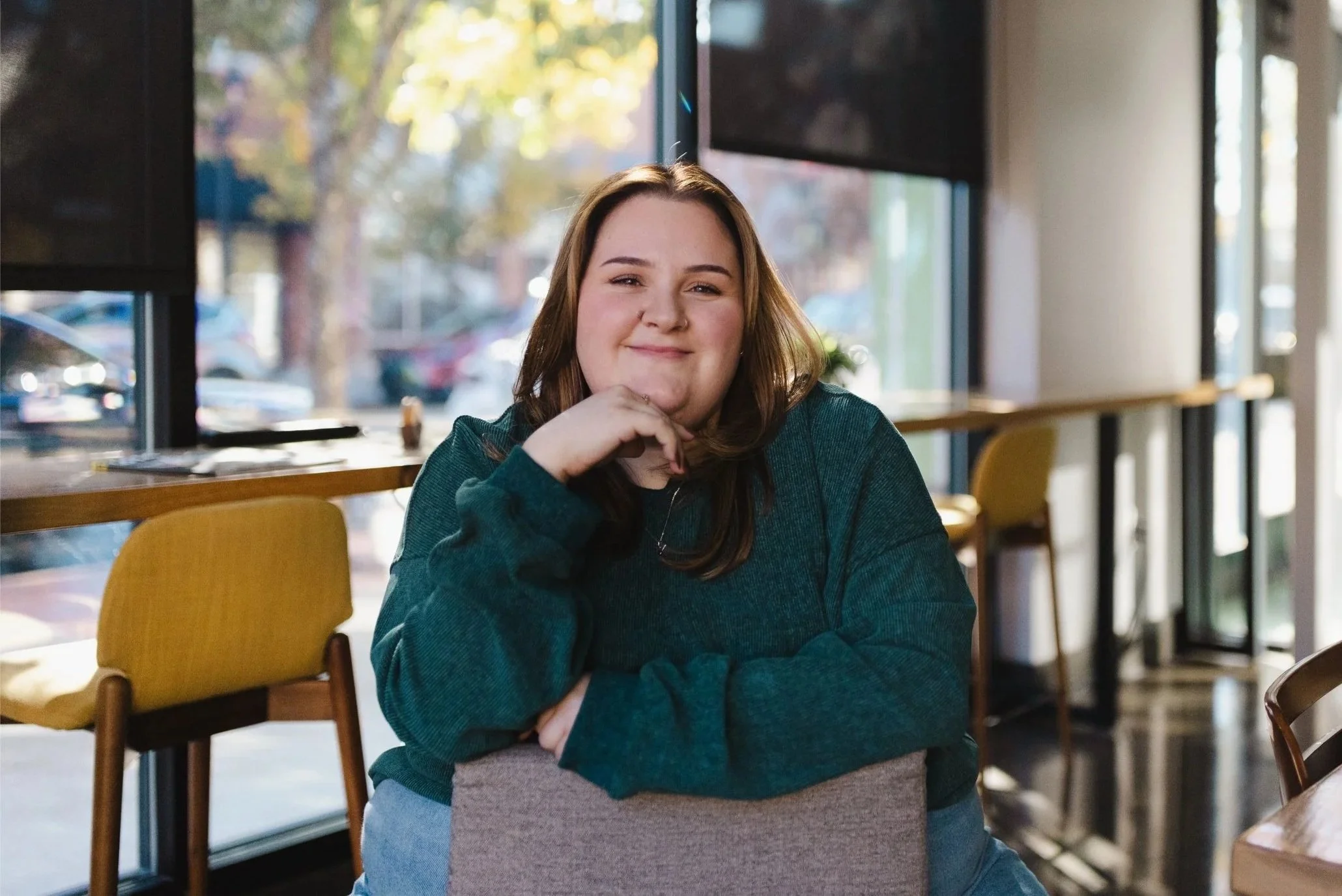 A woman with brown hair smiling, sitting inside a cafe with large windows and outdoor view.