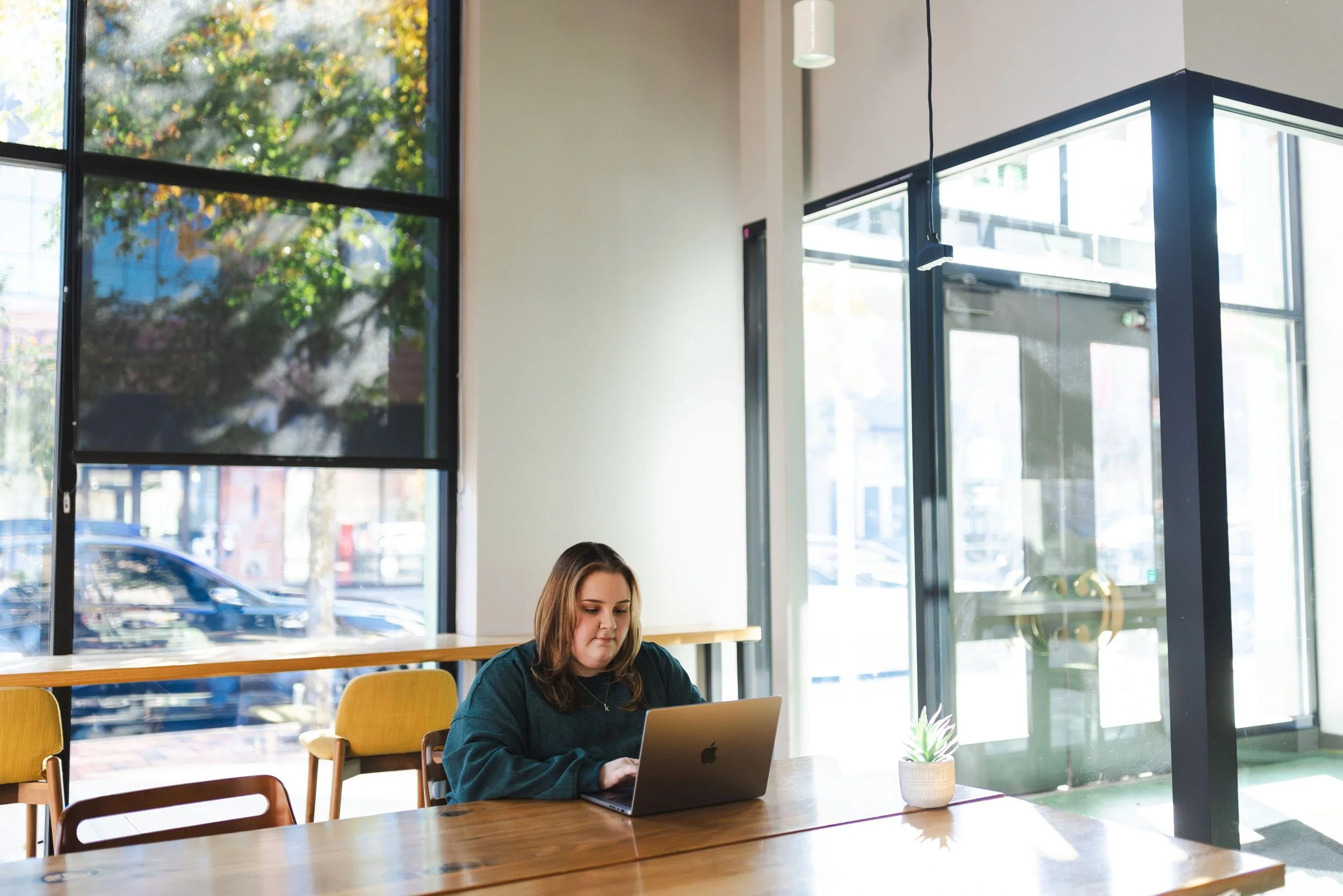 Young woman working on a laptop at a wooden table in a brightly lit cafe with large windows and a small potted plant.