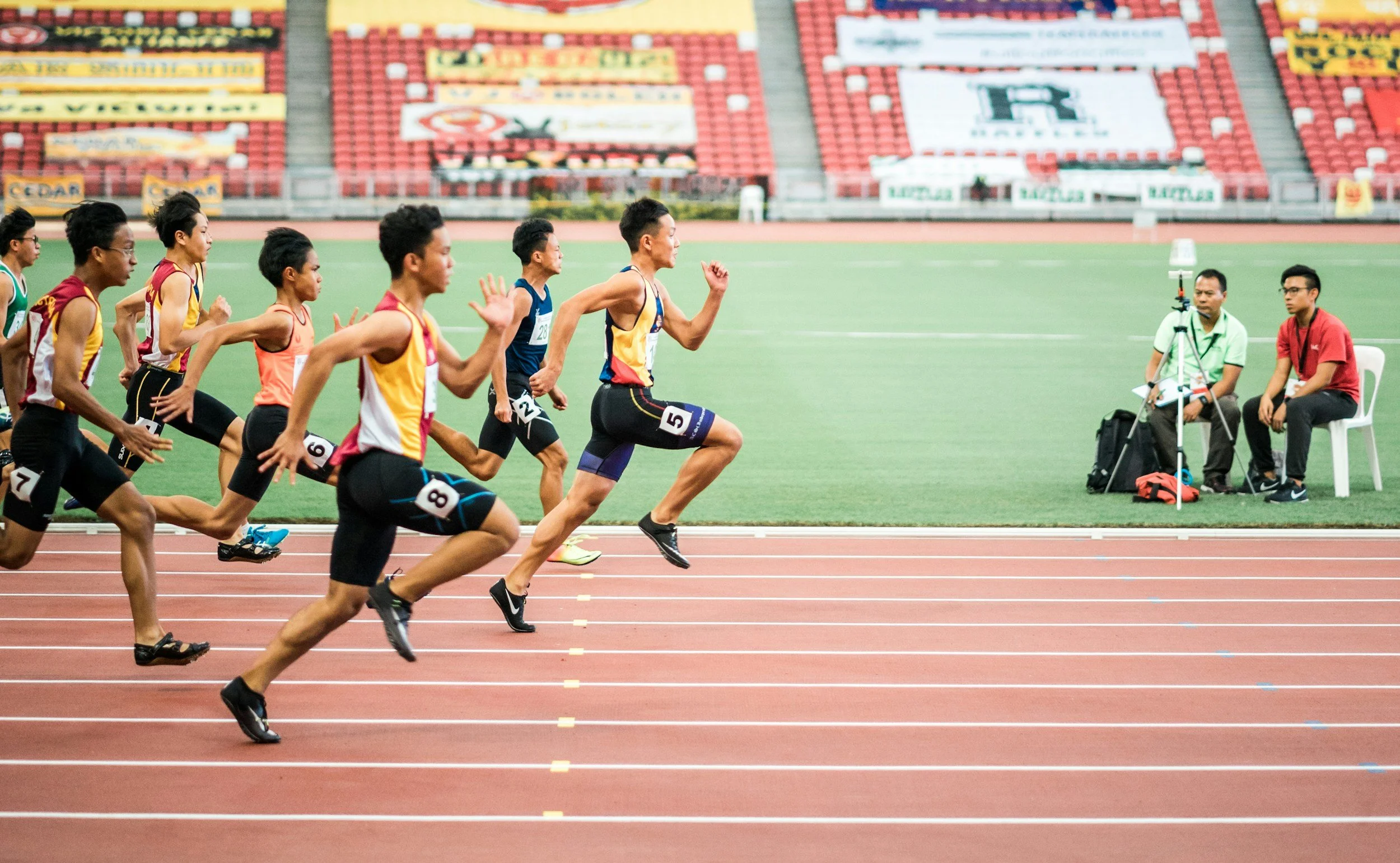 Group of runners competing in a race on a track at a stadium with empty red seats in the background. Two officials sit on chairs beside the track.