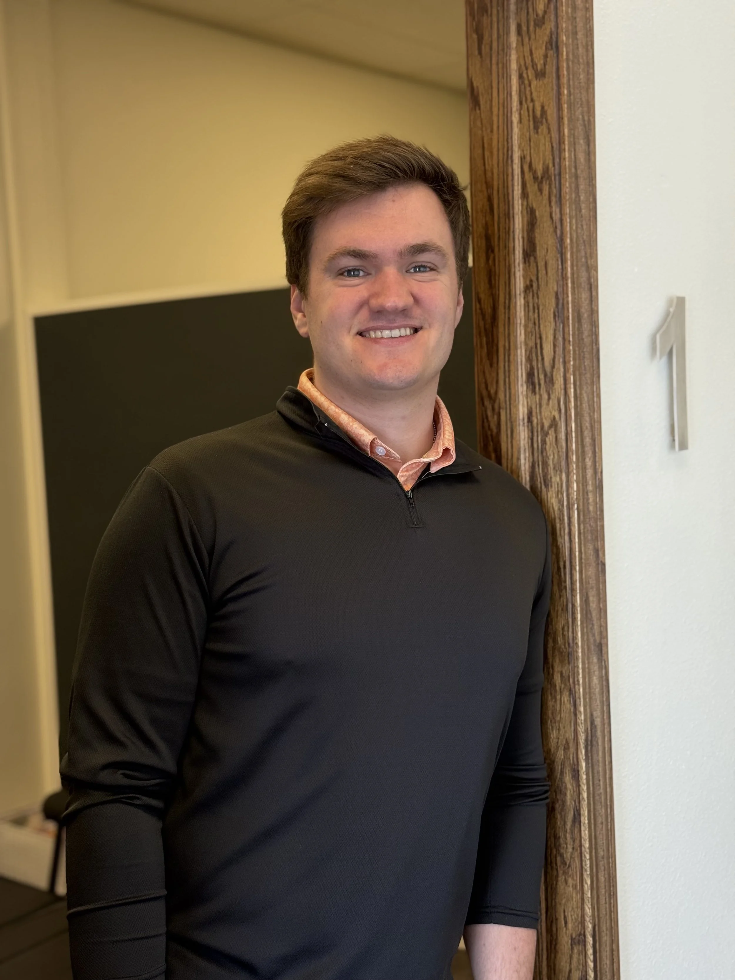A young man with short brown hair, smiling, standing indoors near a wooden door frame, wearing a black quarter-zip pullover and a peach-colored collared shirt.