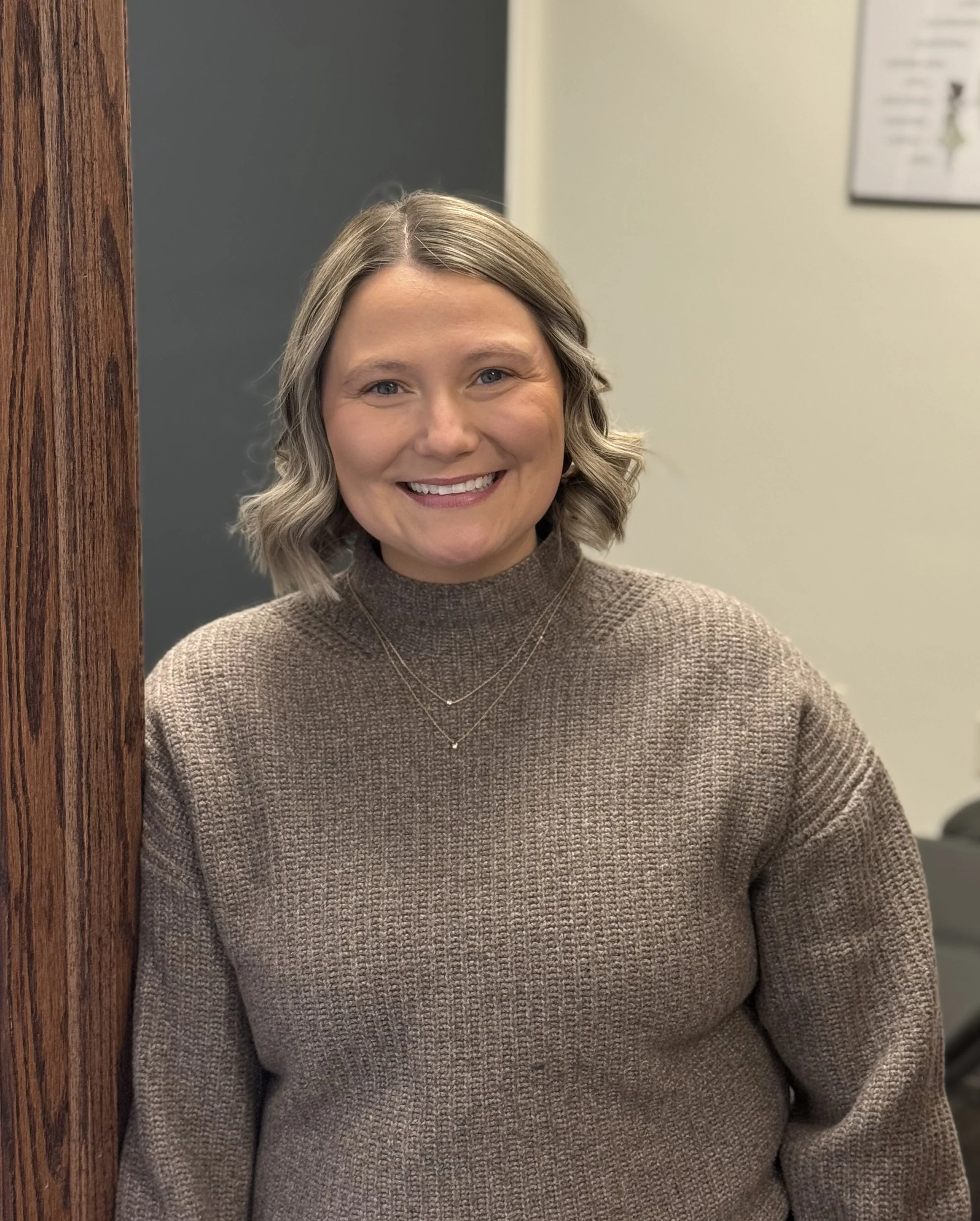 A smiling woman with shoulder-length blonde hair in loose waves, wearing a brown turtleneck sweater and layered necklaces, standing indoors next to a wooden pillar.