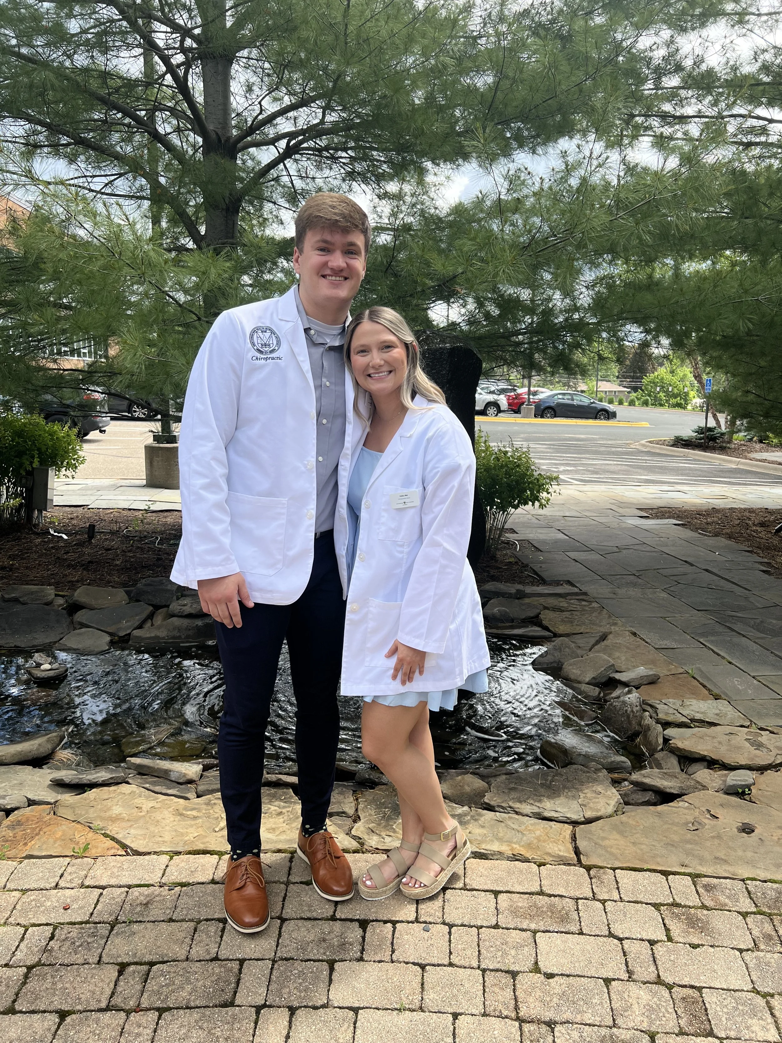 Two young adults, a man and a woman, smiling and dressed in white lab coats, standing outdoors in front of a tree and small pond, with a parking lot in the background.