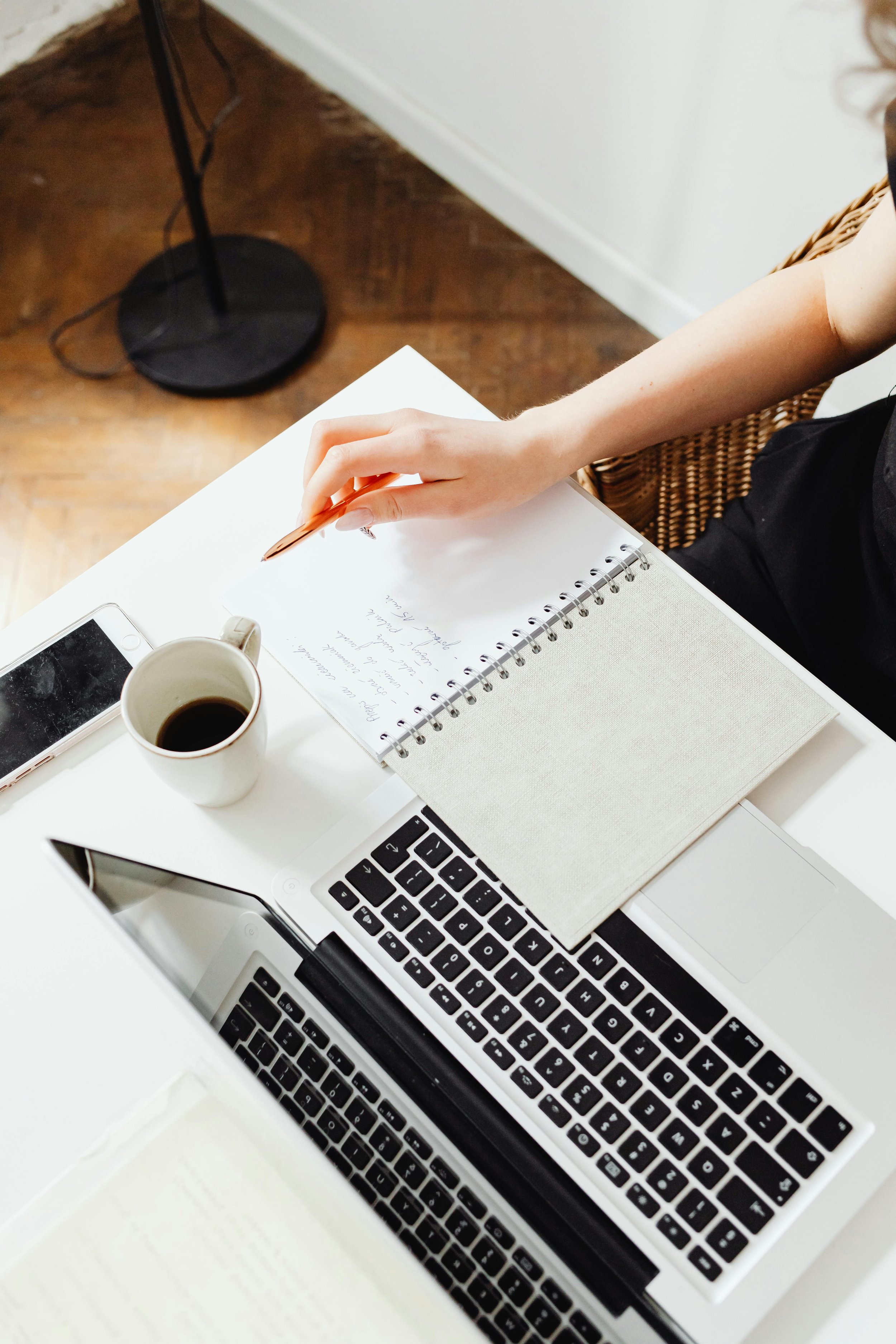 A person sitting at a white desk with a laptop, a smartphone, a notebook, and a cup of coffee, writing with a pen.
