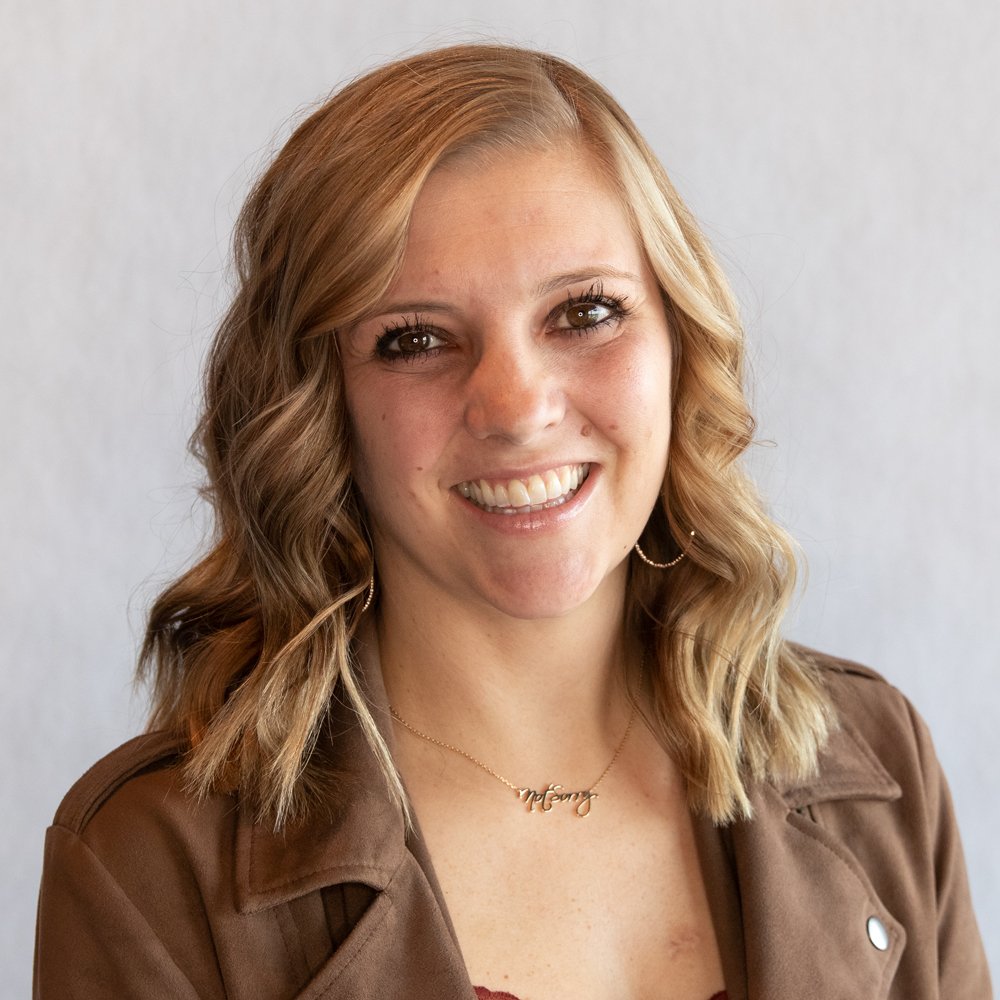 A young woman with wavy blonde hair smiling at the camera, wearing a brown jacket and a necklace that says 'Mommy'.