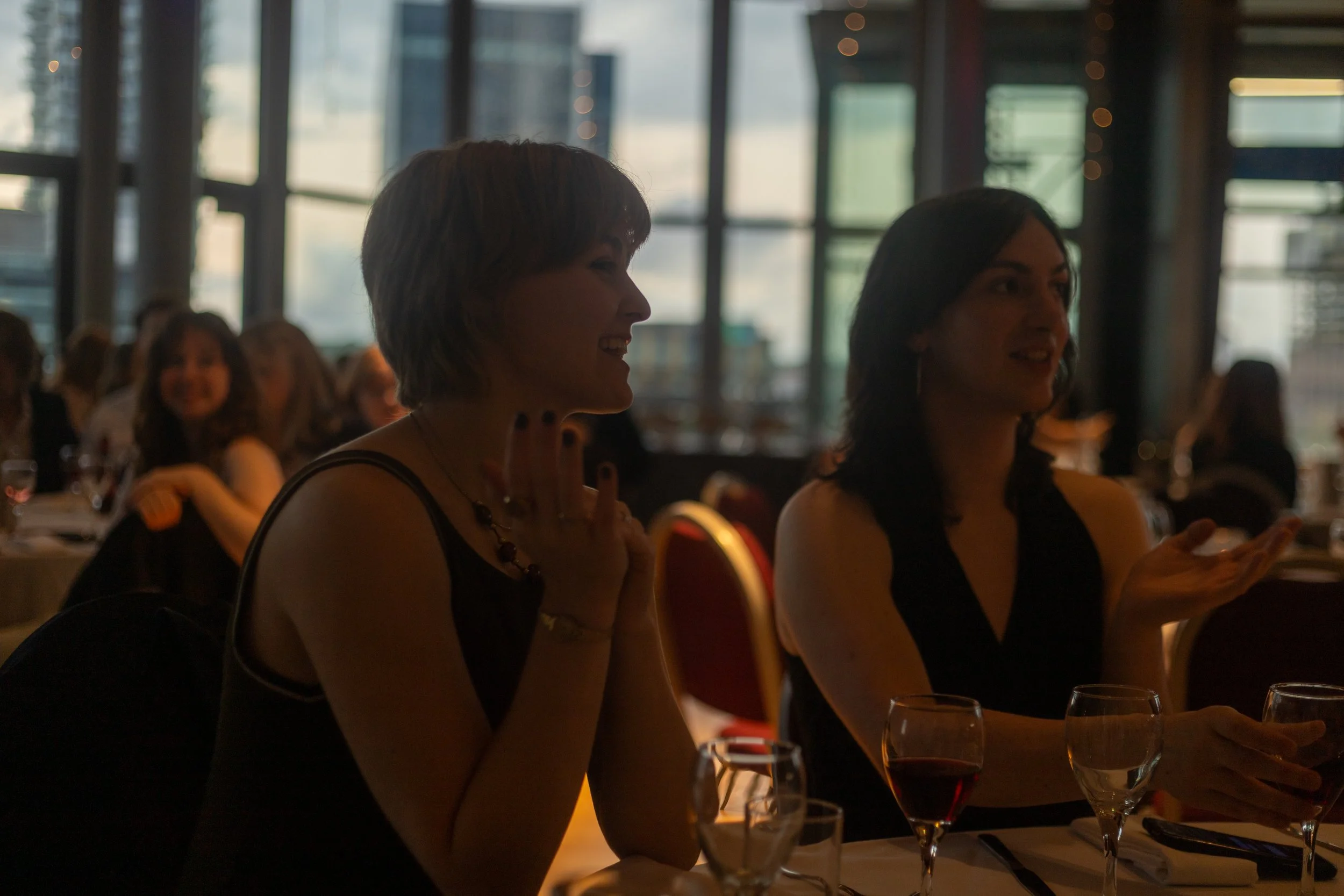 Two women sitting at a dimly lit restaurant or banquet with large windows, engaging in conversation with glasses of wine in front of them.