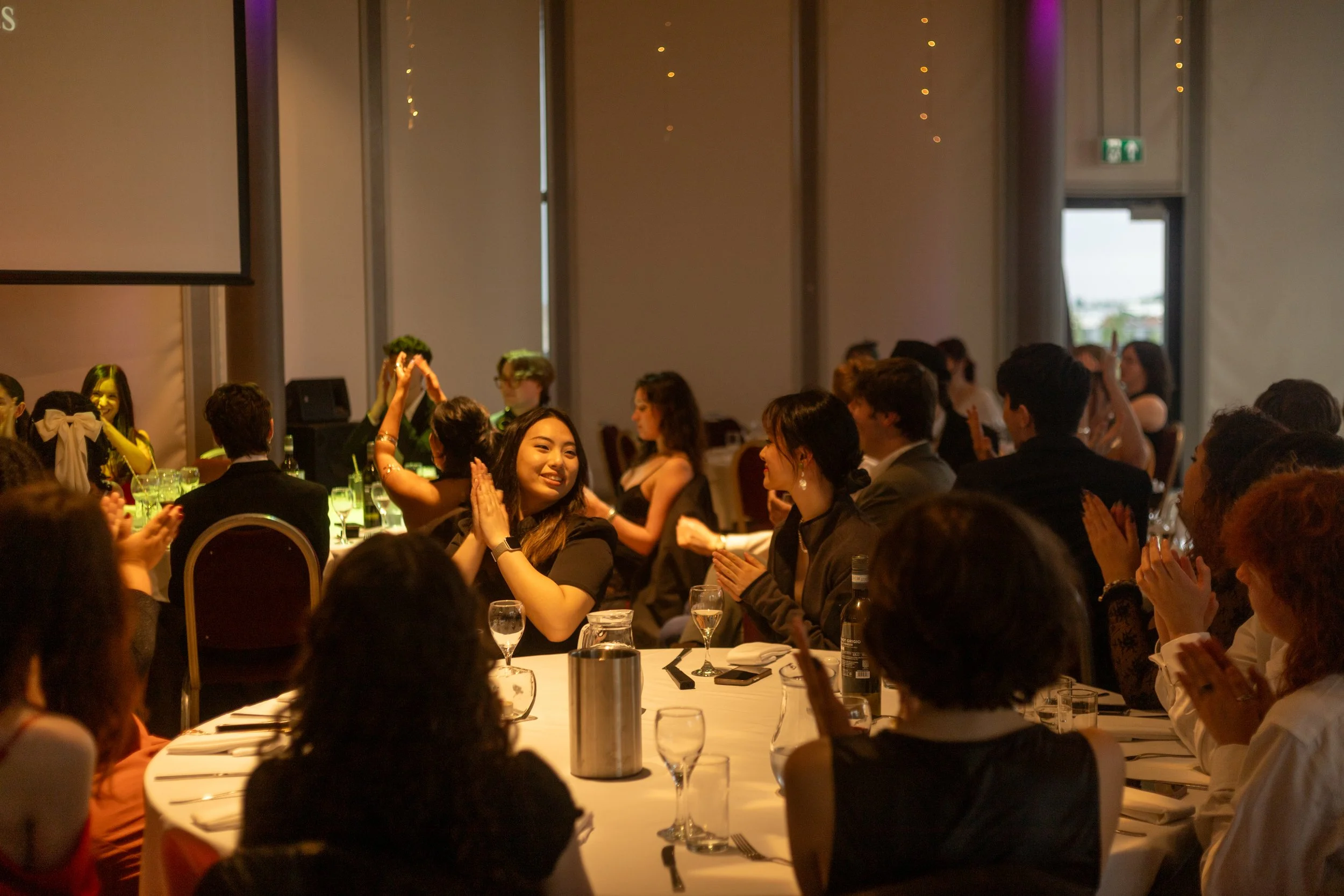 People sitting around a banquet table at a formal event, clapping and smiling, with some raising hands in celebration inside a decorated banquet hall.