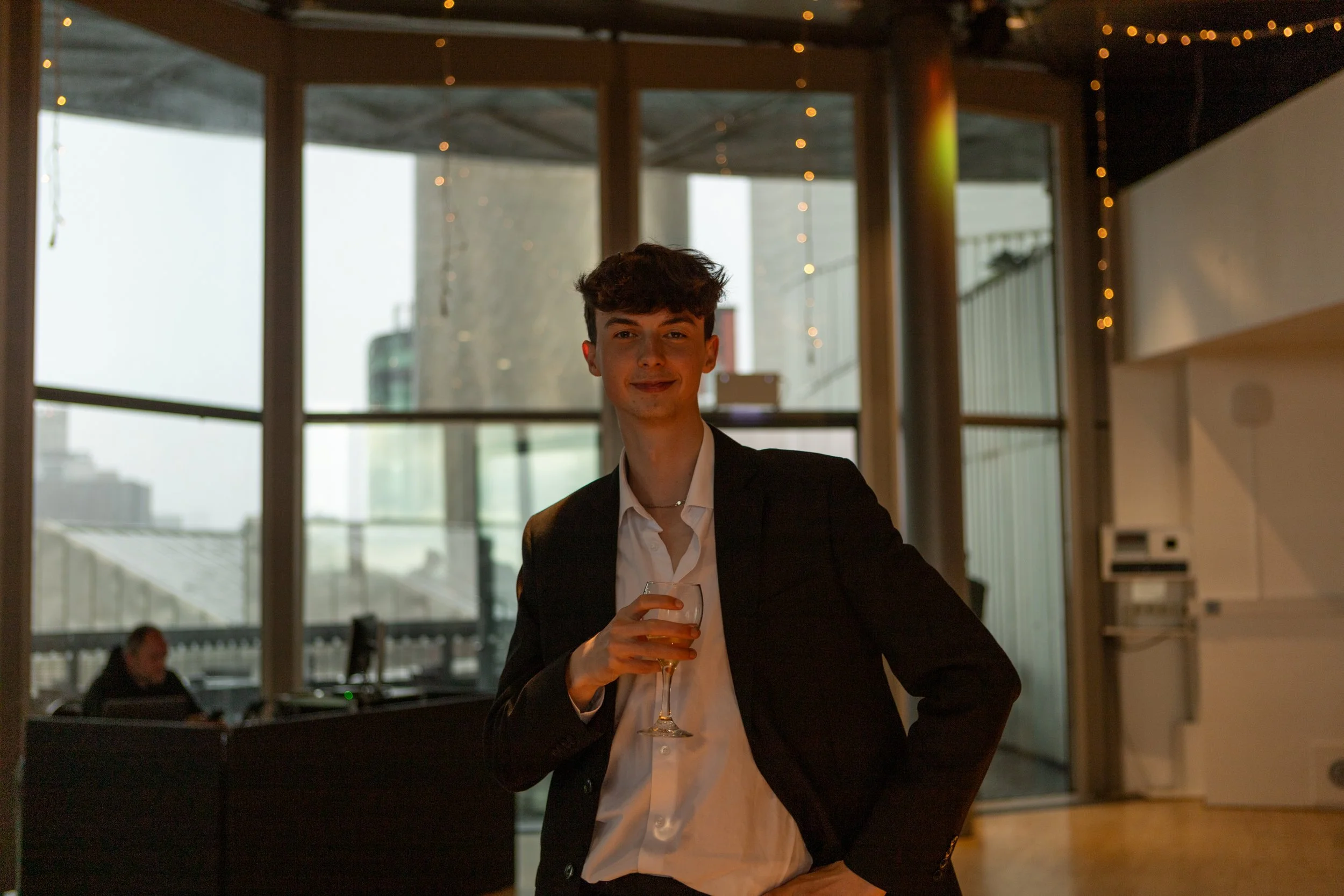Young man in a black suit holding a glass of white wine, standing inside a modern building with large windows and cityscape view.