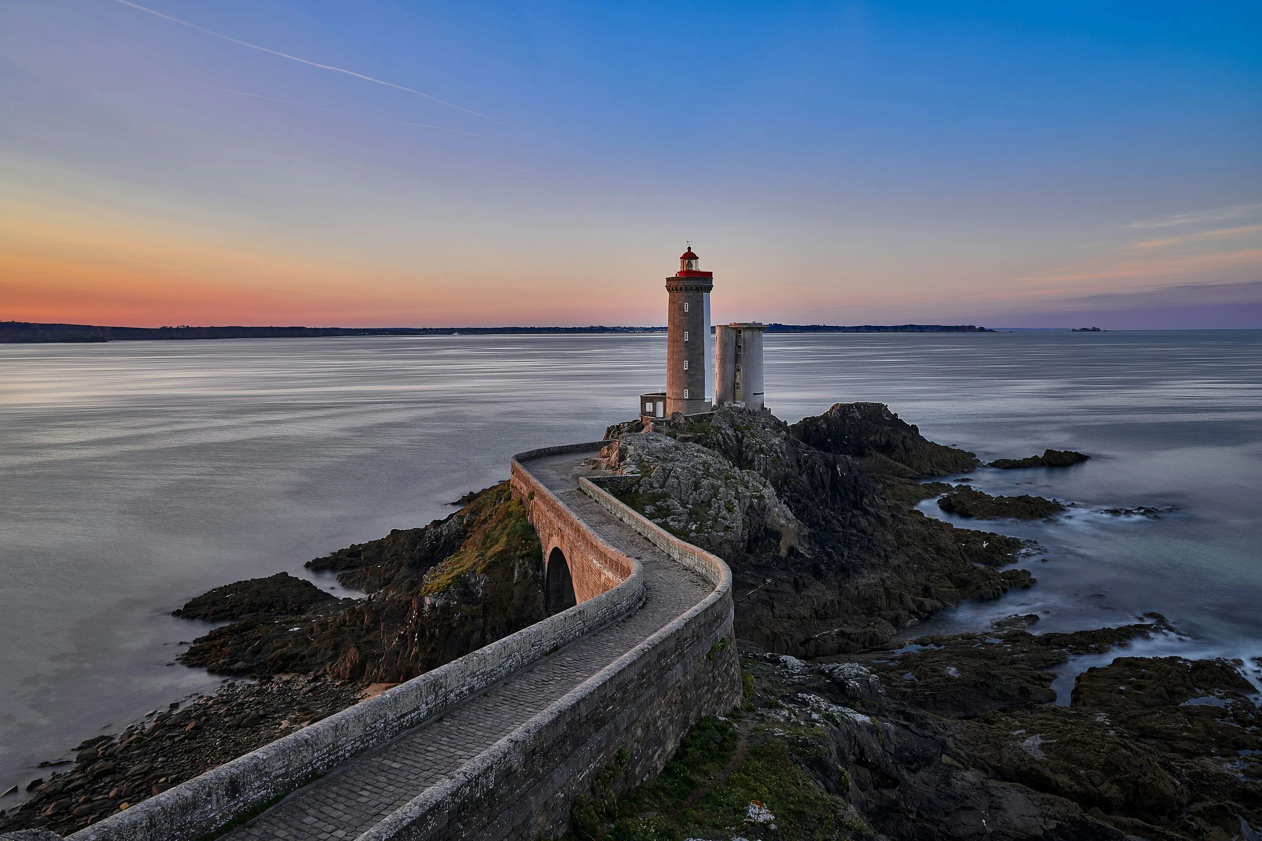 Phare sur une île rocheuse au bord de la mer, avec un chemin sinueux menant au phare, au coucher de soleil.