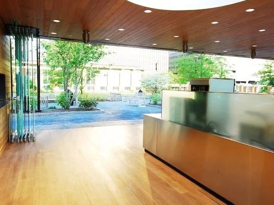 Empty interior space with a wooden floor and a frosted glass partition. Large windows reveal a view of trees and outdoor benches. brahlerdesign.