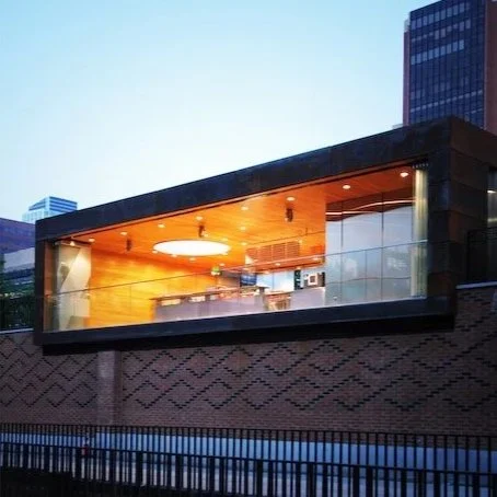 Modern building with a glass balcony and warm interior lighting at dusk. brahlerdesign.
