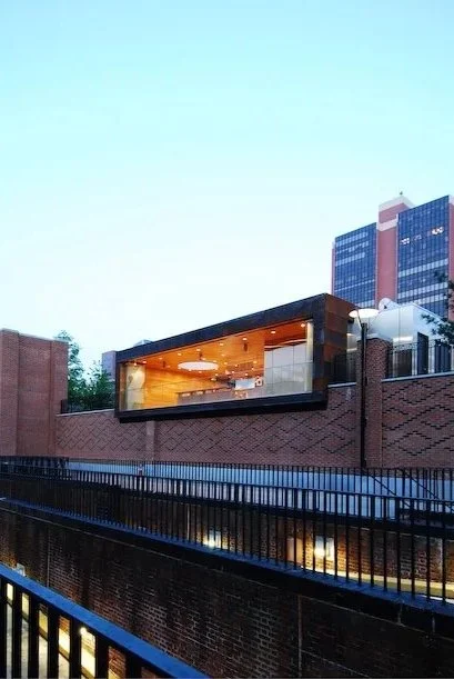 Modern building with large glass windows and brick exterior, with cityscape and tall buildings in the background during evening. brahlerdesign.