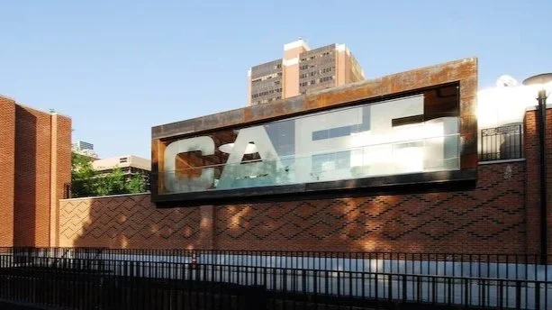 A glass signboard with the word 'CAFÉ' mounted on a brick wall under a clear blue sky. brahlerdesign.
