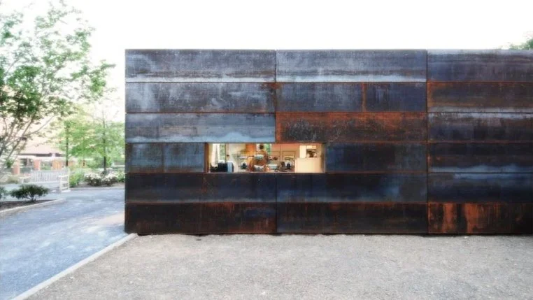 Modern building facade made of large dark metal panels with a horizontal window showing a glimpse of the interior. brahlerdesign.