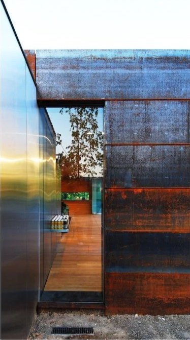 Modern architectural view showing a narrow alleyway between reflective metal and rusted metal walls with a glimpse of trees and sky in the background. brahlerdesign.