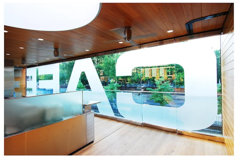 Interior view looking out through large frosted glass windows with partial company logo visible, showing a modern space with wooden ceiling and a city street view outside. brahlerdesign.