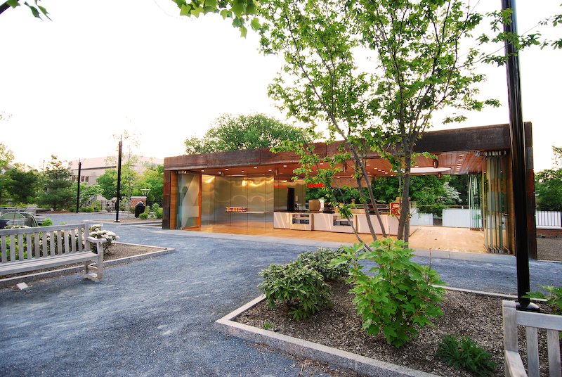 Modern building with glass storefront, outdoor seating area, trees, and a gravel pathway. brahlerdesign.