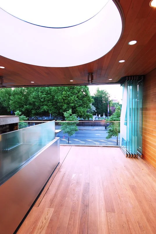 Modern balcony with wooden flooring, glass railing, and a large circular light fixture on the ceiling, overlooking green trees and a street. brahlerdesign.