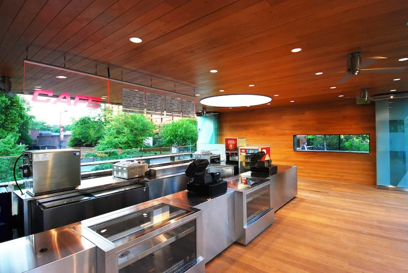 Modern cafe counter with stainless steel equipment, cash register, menu board, and large window showing trees outside in a wooden interior. brahlerdesign.