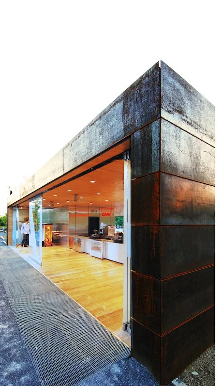 Exterior of a modern building with dark metal panels and a large open window showing a food counter inside, with wooden flooring and a person in a white coat nearby. brahlerdesign.
