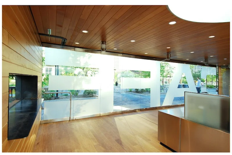 Empty room with large glass windows, wooden ceiling and floor, and some outdoor greenery visible through the windows.