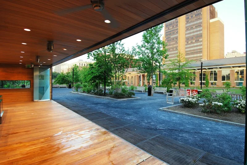 View of a modern outdoor park area with trees, benches, and a sidewalk, seen from inside a building with wooden flooring and ceiling. brahlerdesign.