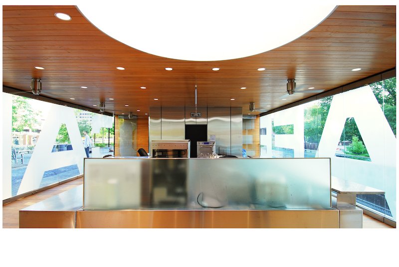Interior of a modern fast-food restaurant with a wooden ceiling, stainless steel counter, and large windows with partial white lettering spelling 'A' and 'L' on the glass.