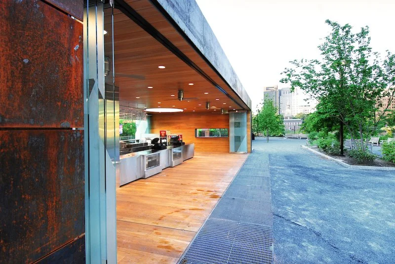 Outdoor area with stainless steel kitchen appliances and oven, adjacent to a modern building with a wood-paneled roof, overlooking trees and cityscape. brahlerdesign.
