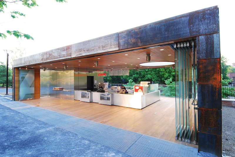 Modern open-concept cafe with stainless steel counter, wooden ceiling, and rusted metal exterior, surrounded by trees. brahlerdesign.