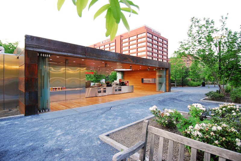 Modern outdoor café with a sleek, minimalist design, featuring a stainless steel counter, served by an attached building with a wooden and glass façade, surrounded by greenery and trees in an urban park setting. brahlerdesign.