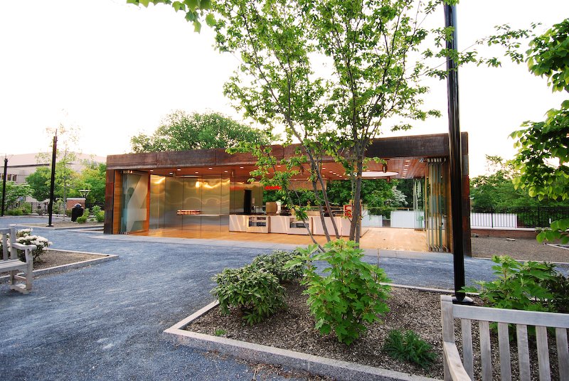 Modern glass building with wooden accents, surrounded by trees and greenery in an outdoor park setting during daytime. brahlerdesign.