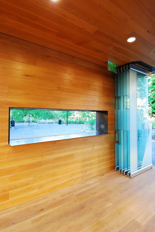 Interior of a building with wooden walls and floor, a rectangular window looking outside to trees and a pathway, and glass doors with vertical blinds near an exit sign. brahlerdesign.