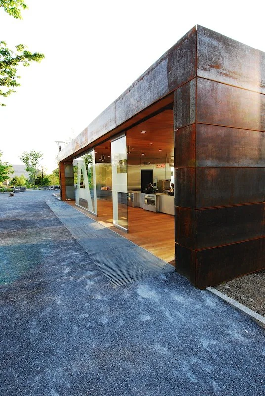 Modern building with large glass windows, rust-colored metal exterior, and a visible interior kitchen area, located in a parking lot with trees in the background. brahlerdesign.