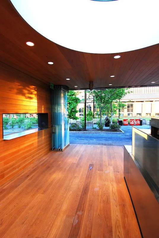 Interior view of a modern establishment with wooden flooring and walls, a curved wooden ceiling with installed lights, large open entrance with glass doors, outside view of trees and buildings, and Coca-Cola coolers in the background. brahlerdesign.