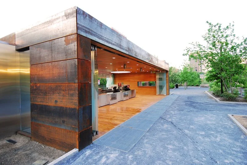 Modern outdoor concession stand or food kiosk with a rusty metal exterior, open windows, and wood interior, located in a park or plaza with trees and pathways. brahlerdesign.