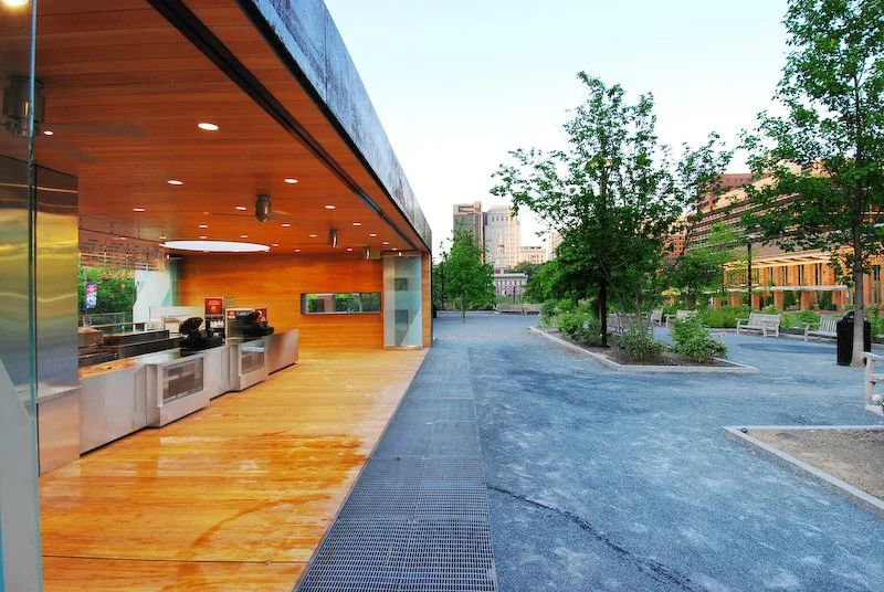 Empty outdoor park with a modern ice cream or snack stand, trees, benches, and city buildings in the background during daytime. brahlerdesign.
