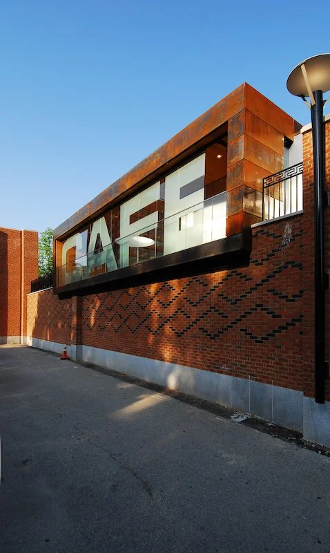 Modern building with large glass windows and rust-colored metal accents, part of a brick structure, under a clear blue sky. brahlerdesign.
