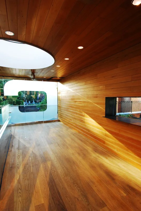 Empty modern room with wooden walls and ceiling, large circular window, and floor-to-ceiling glass door leading outside, sunlight casting shadows inside. brahlerdesign.