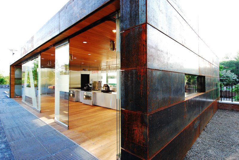 Modern cafe with glass exterior, wooden ceiling, and stainless steel counter, surrounded by rusted metal panels, on a sunny day. brahlerdesign.