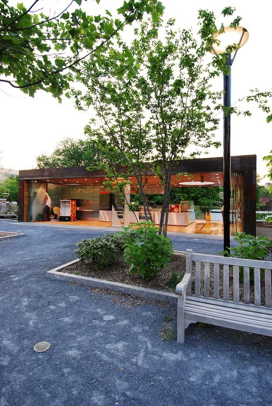 A small outdoor food stand with a modern design, surrounded by trees and greenery, a bench, and a streetlamp nearby. brahlerdesign.