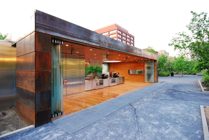 Modern outdoor food stand with stainless steel kitchen equipment, wooden ceiling, and wood flooring, situated in a park with trees and a blue paved walkway. brahlerdesign.