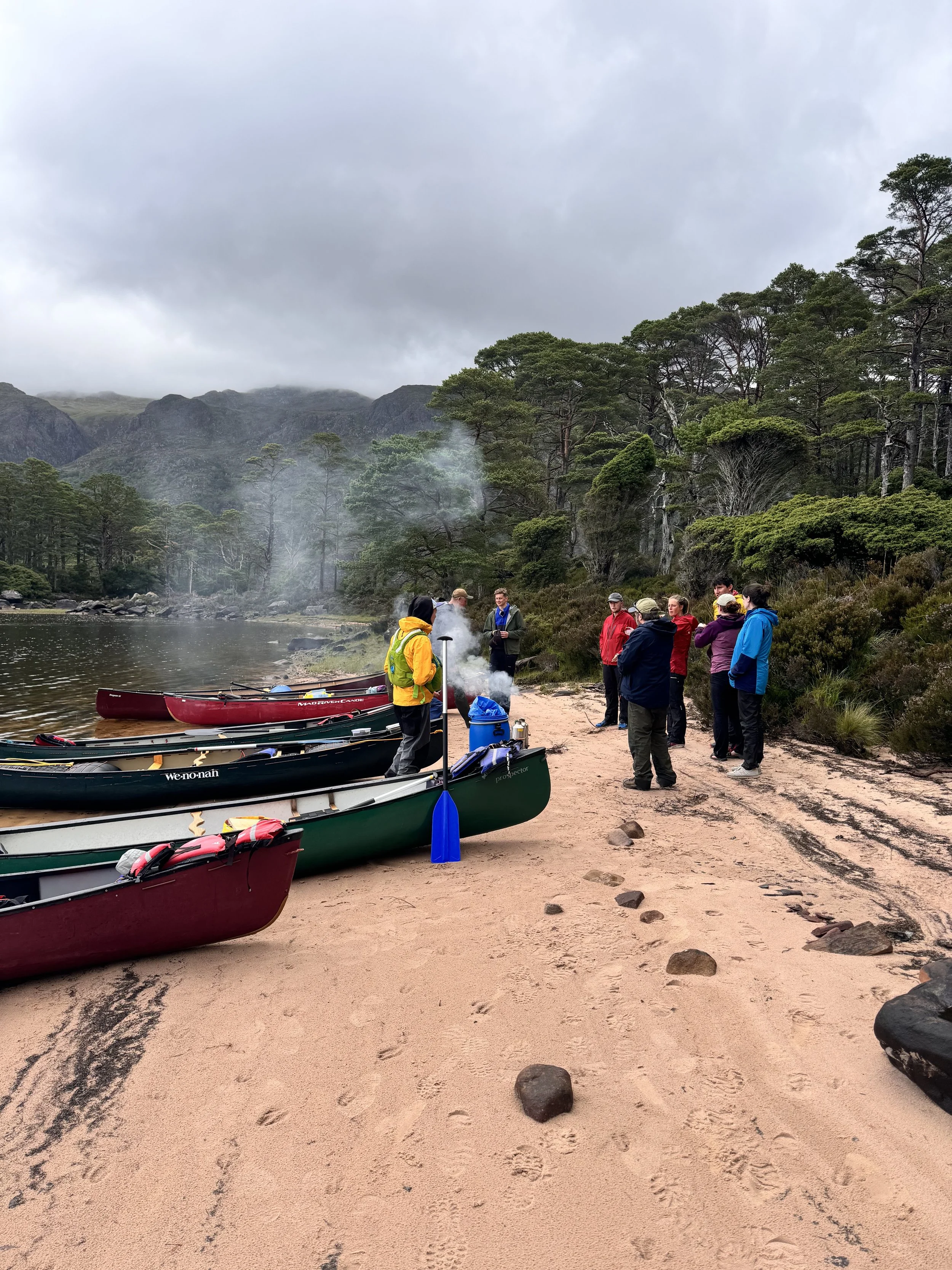 Group of people by canoes on a sandy lakeshore, surrounded by trees and mountains, with cloudy sky and smoke or steam in the air