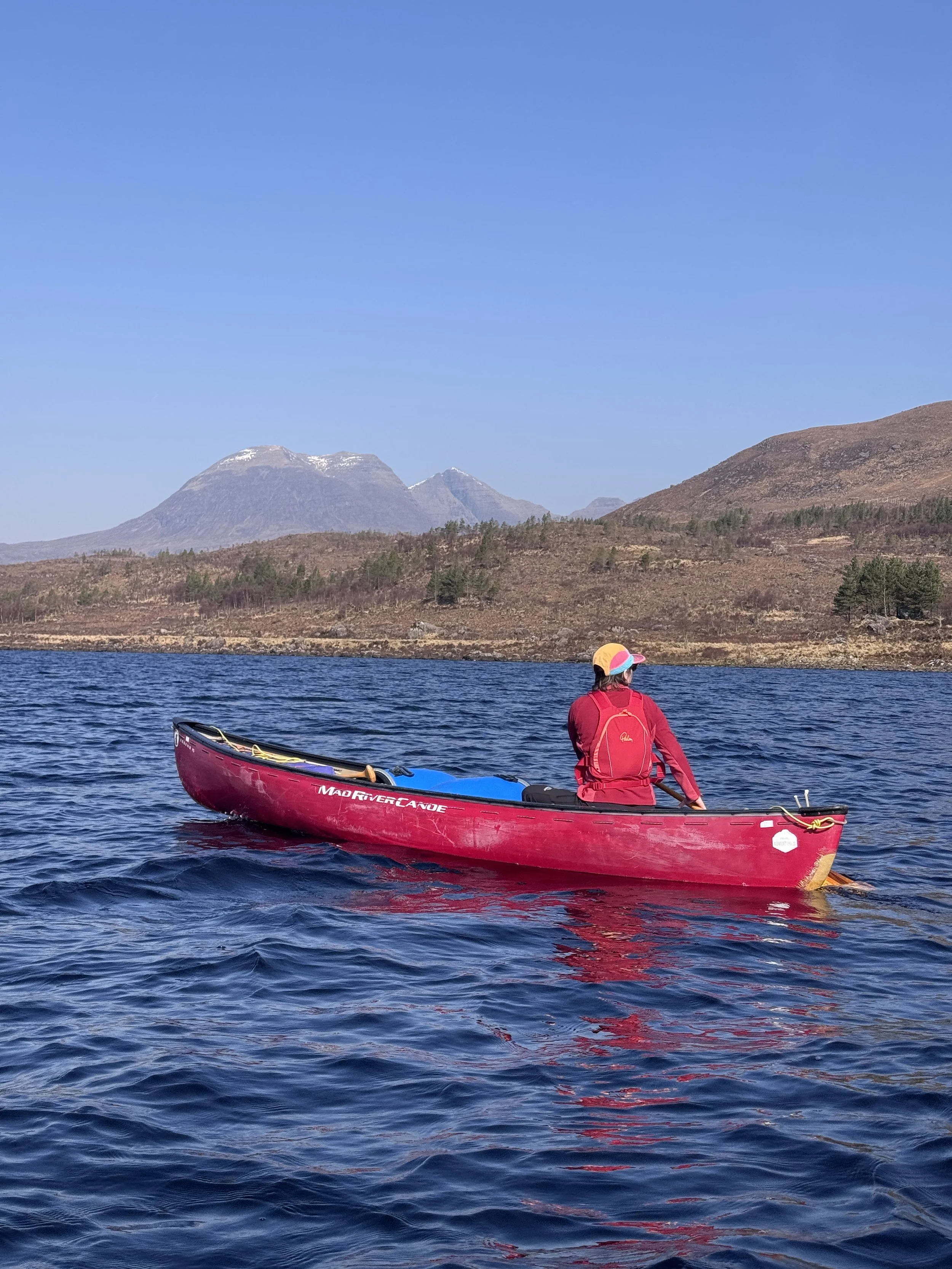 Person in a red kayak on a body of water with mountains in the background under a clear blue sky.