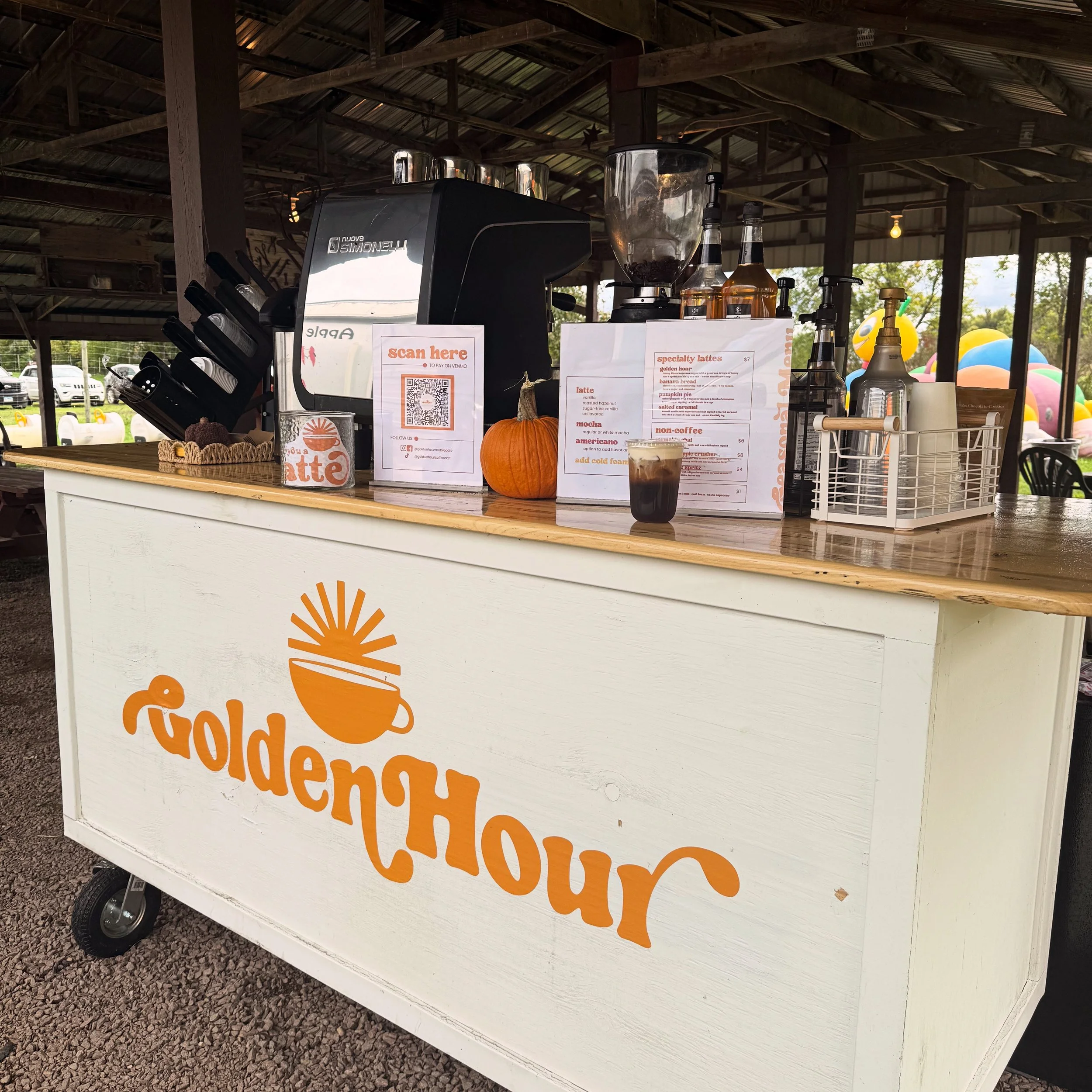 A mobile coffee stand with the name 'Golden Hour' displayed on the front in orange lettering and an illustration of a steaming cup and sun. The stand has a wooden countertop with a small pumpkin, a coffee machine, a menu sign, and various bottles. There are balloons in the background and a canopy overhead.