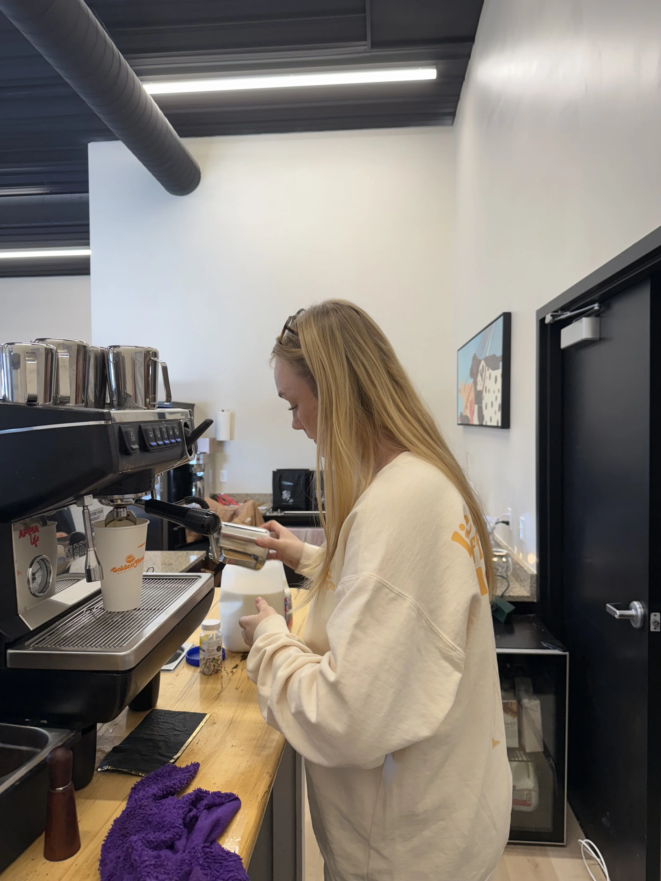 A woman with long blonde hair, wearing a beige sweatshirt, is preparing coffee at a coffee station. There are coffee mugs, a coffee machine, and various supplies on the wooden counter.