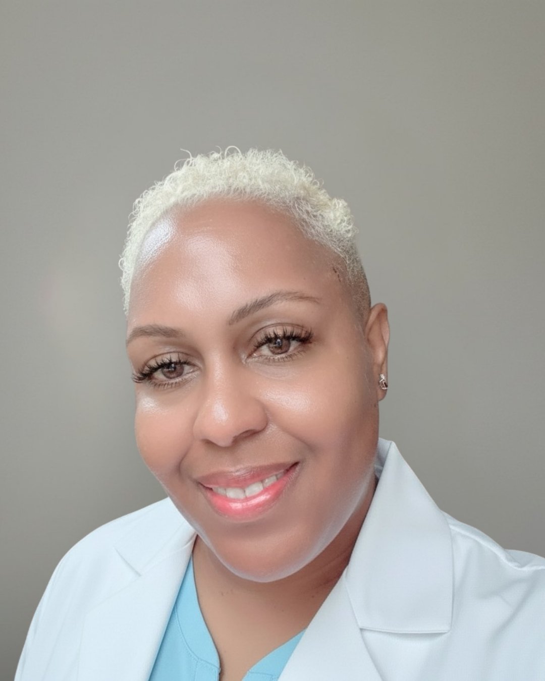 Close-up of Nurse Practitioner Maria Gaines-Onwukwe with short platinum blonde curly hair, smiling, wearing light blue medical scrubs and small earrings, with a plain gray background.