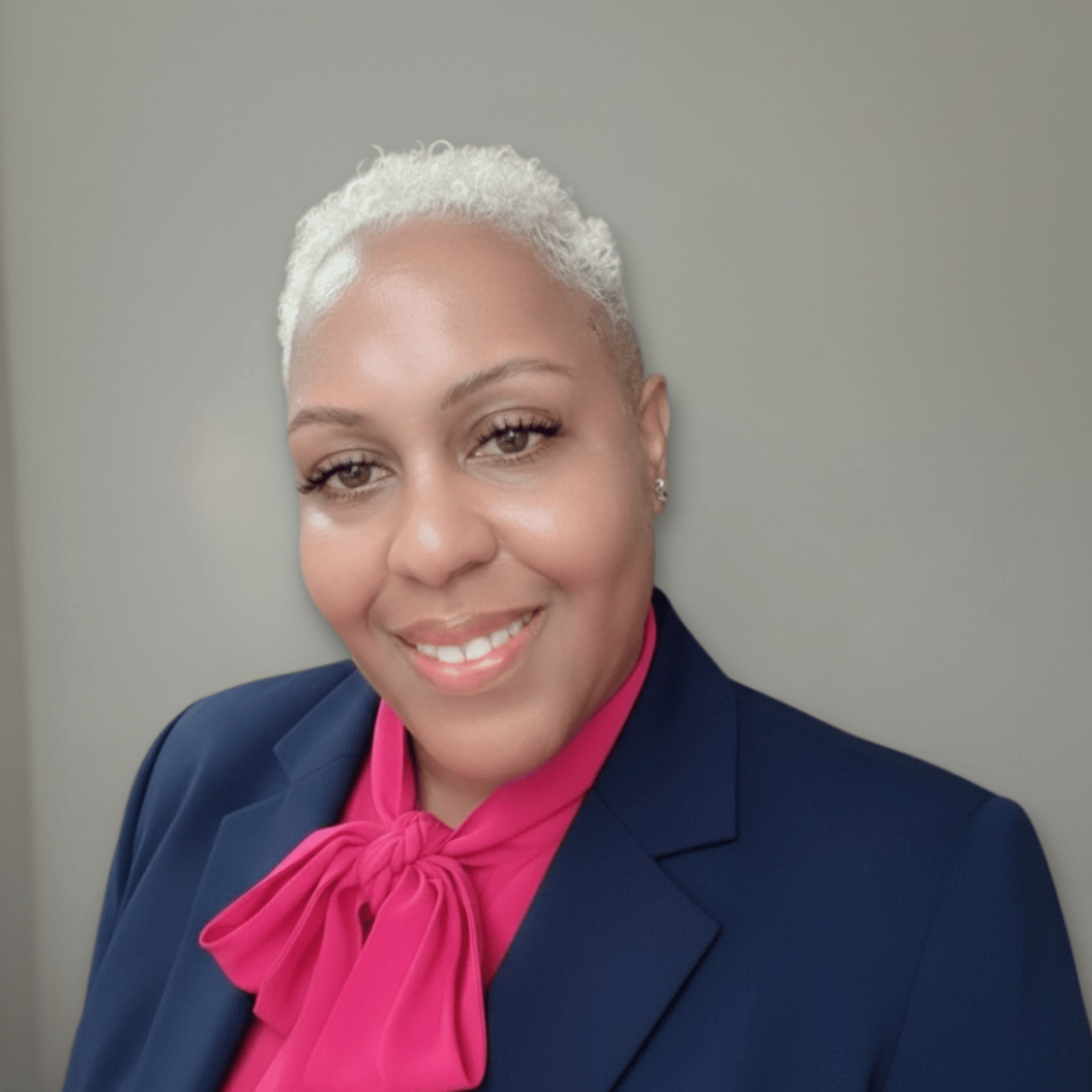 Close-up of a woman with short curly grey hair, wearing a navy blazer and pink blouse with a bow tie, smiling and looking at the camera.