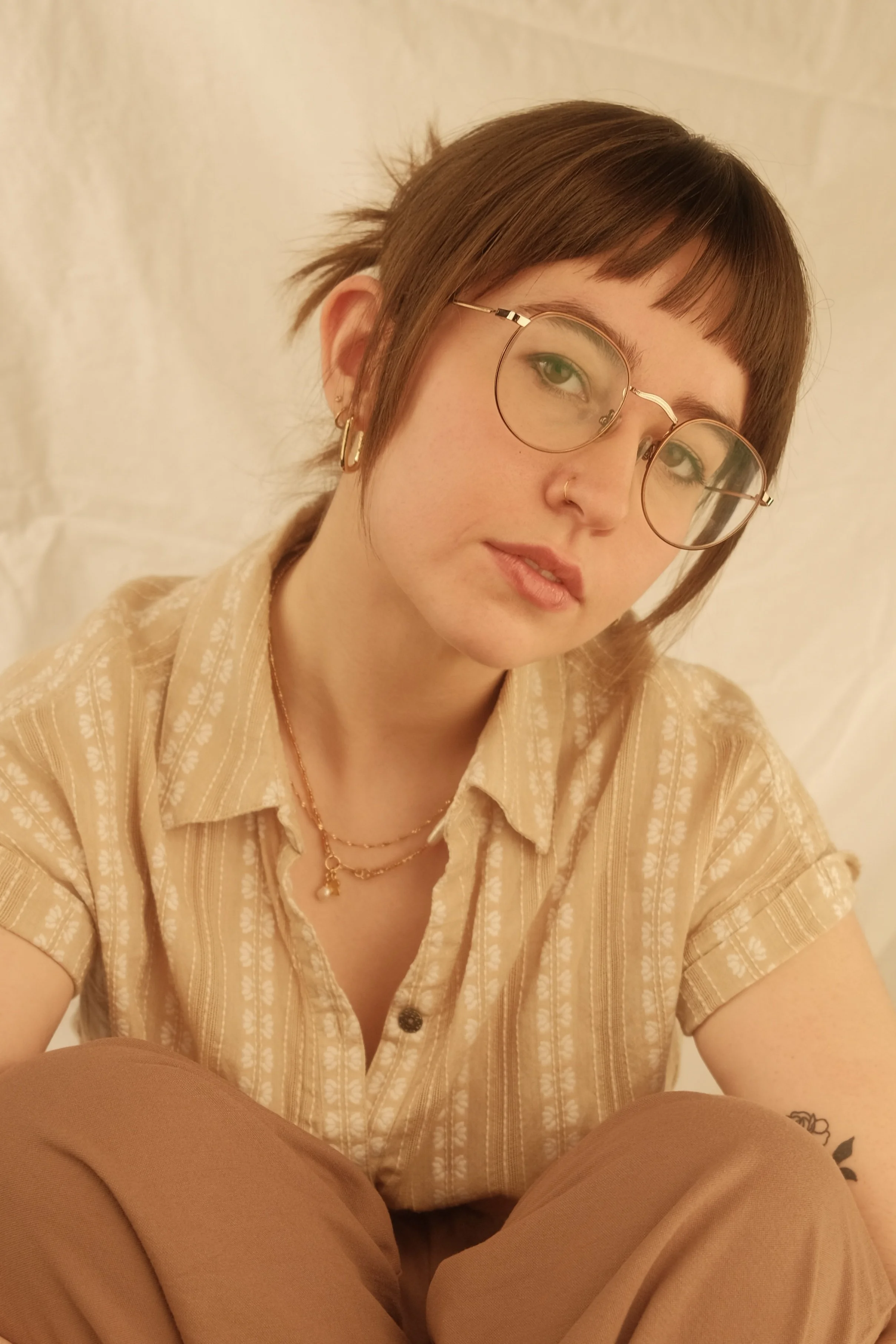A young woman with short brown hair, glasses, and earrings, wearing a beige striped shirt and layered necklaces, sitting against a light-colored background.